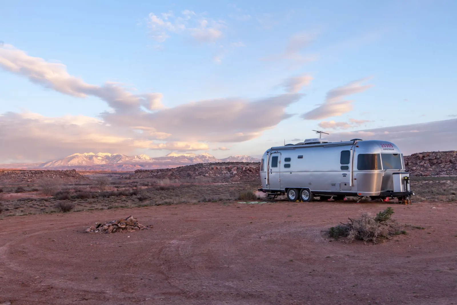 A silver travel trailer parked alone in a desert landscape at sunset with distant mountains