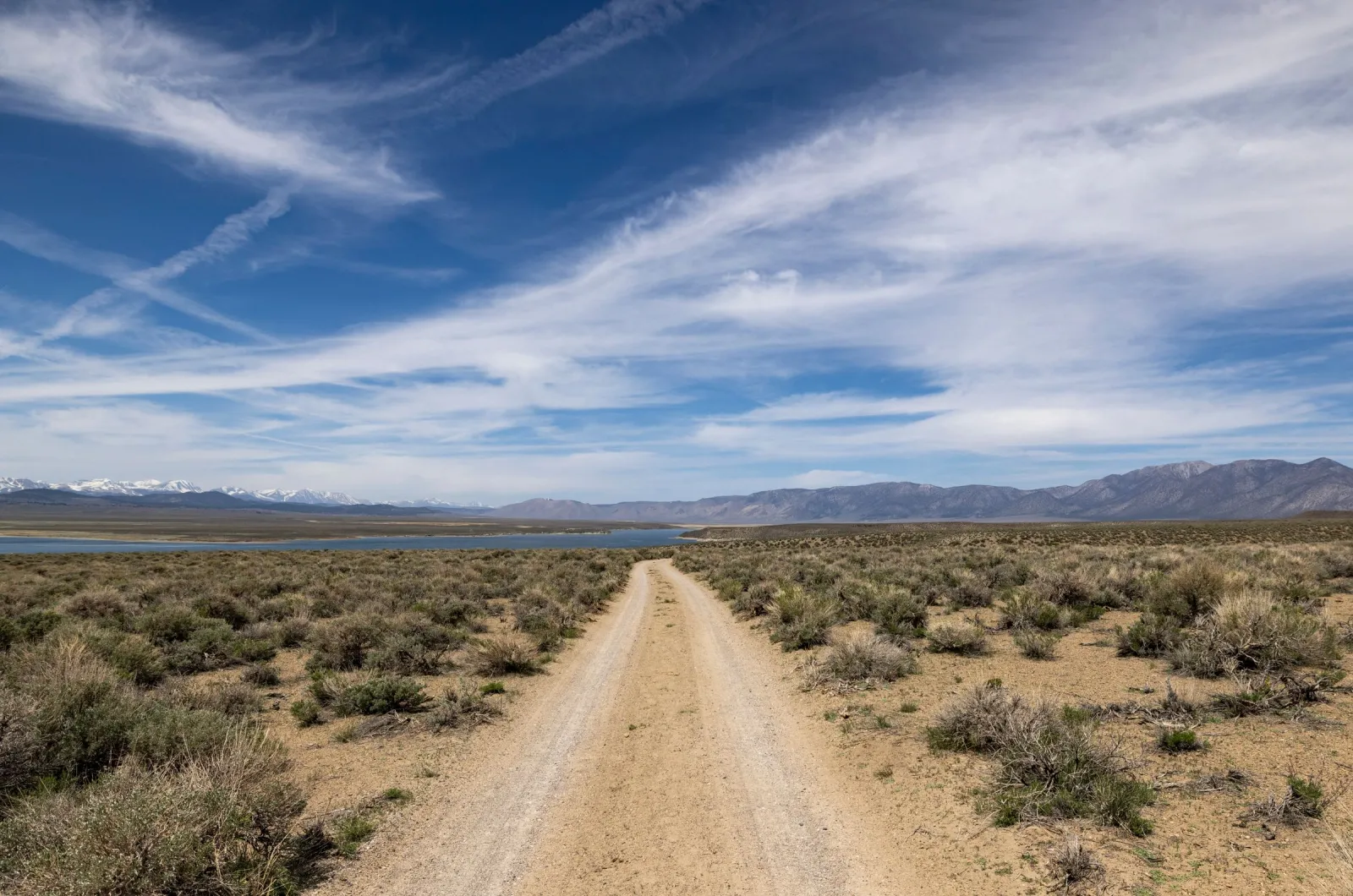 A sandy two-track running through sagebrush toward a lake and distant snowy peaks